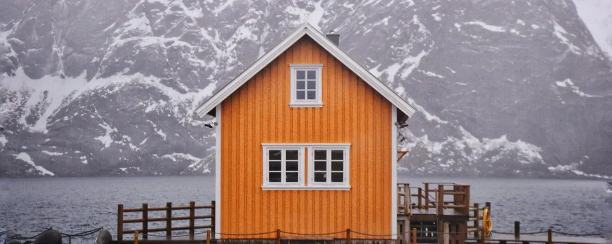 Oransje trehus med saltak på brygge foran snødekte fjell i Lofoten
